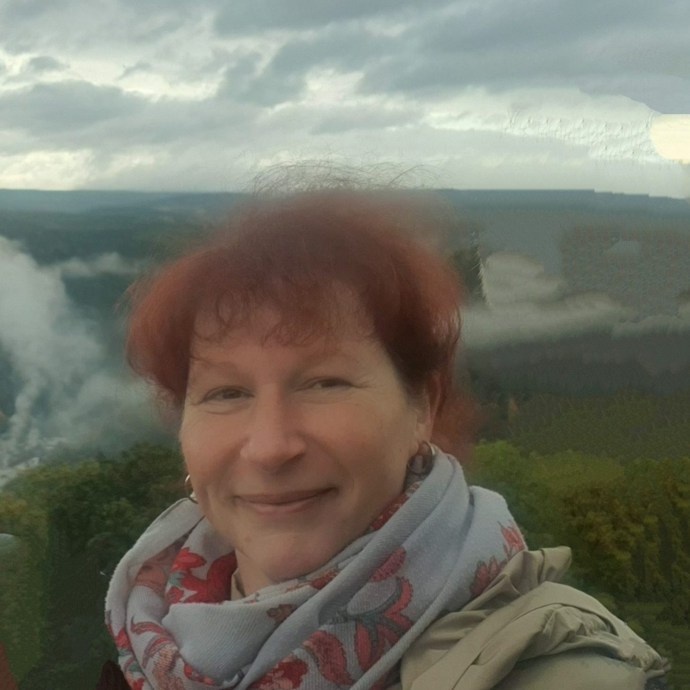 Frau mit rotem Haar, lächelt vor einer wolkigen Naturkulisse.